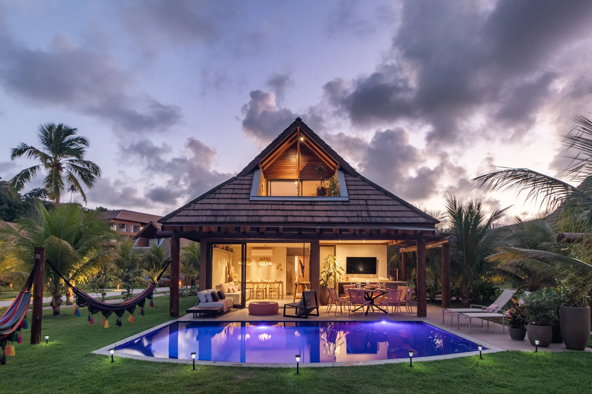 Vista crepuscular completa da fachada chalé do Bangalô 48, com piscina privativa iluminada em azul, deck e terraço gourmet acolhedores