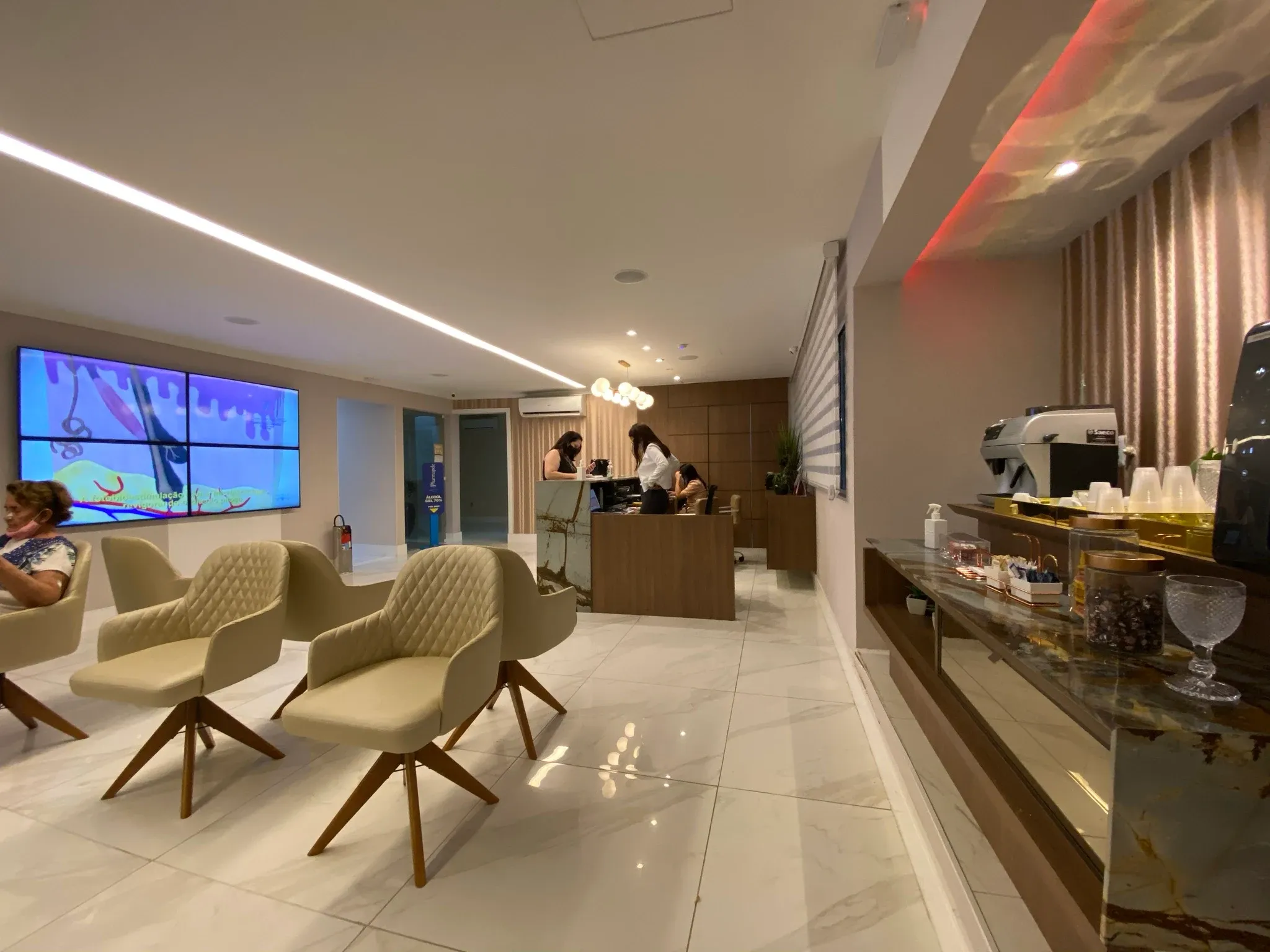 Recepção e sala de espera da clínica com videowall informativo, poltronas giratórias em couro bege matelassê, balcão em madeira e mármore verde, e cantinho de café gourmet