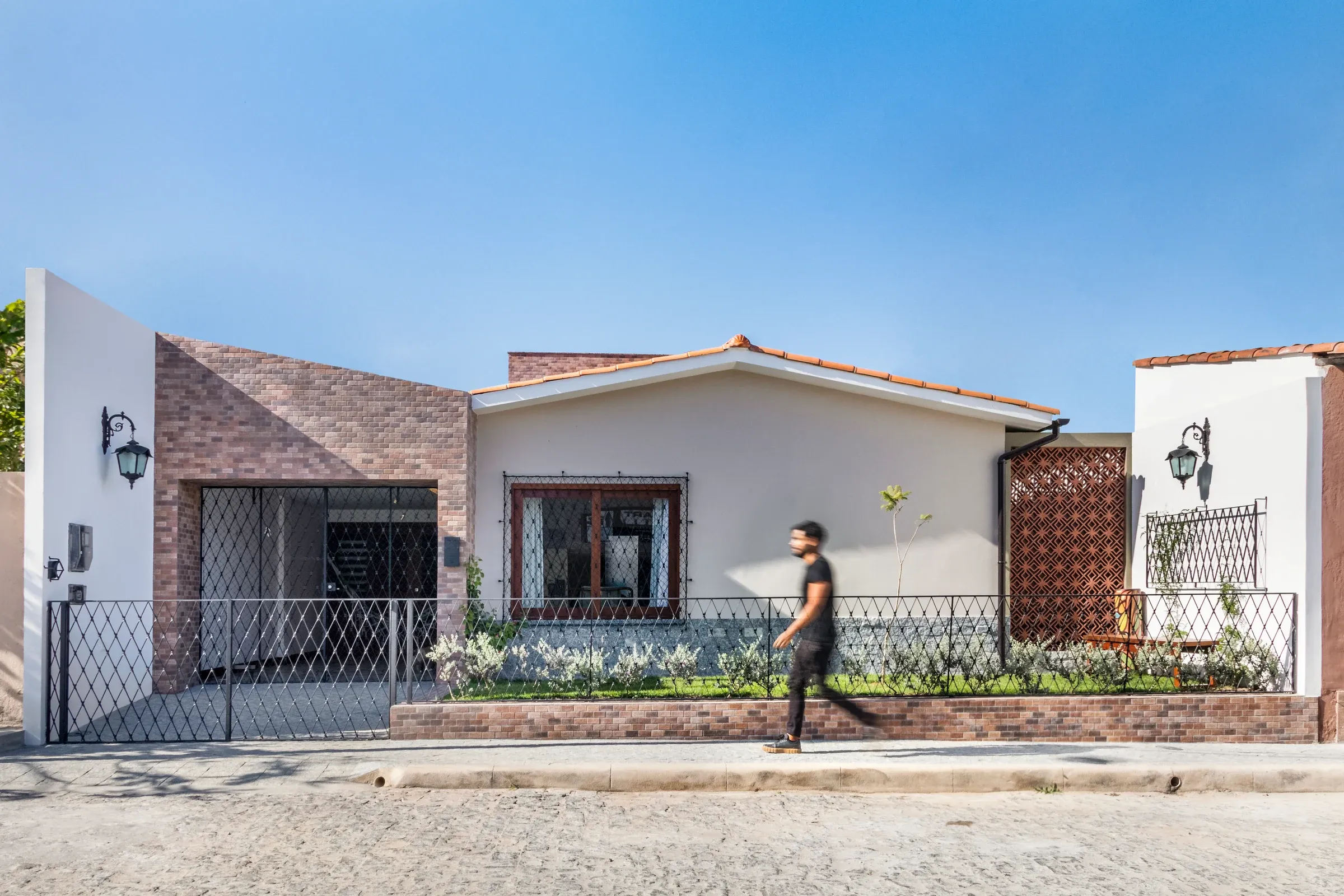 Fachada frontal urbana da casa na rua em paralelepípedos, com volume de entrada em tijolinho aparente, portão geométrico em ferro e cobogós