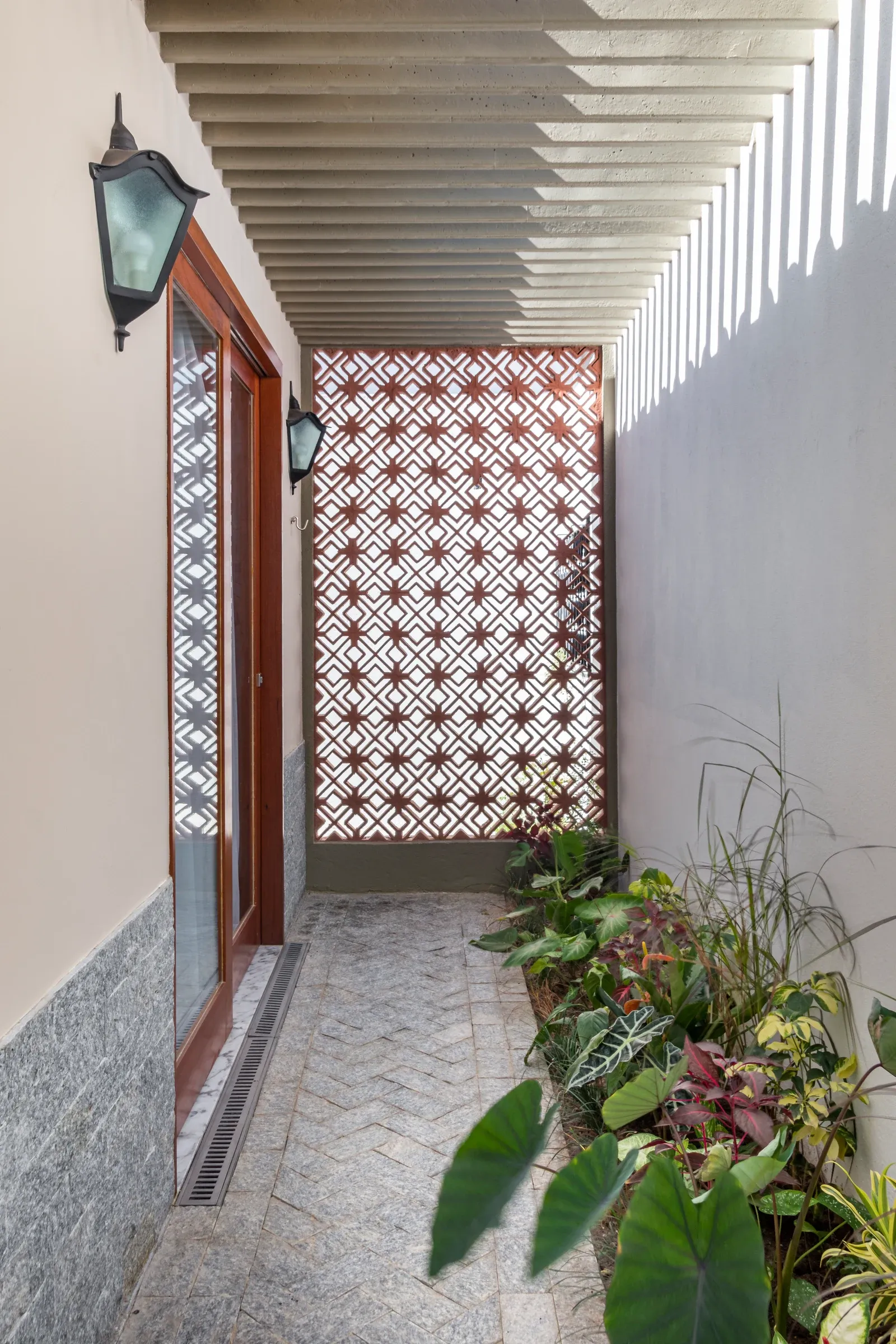 Corredor lateral com pérgula em concreto gerando sombras no piso de pedra, canteiro ajardinado e painel de cobogó cerâmico vazado