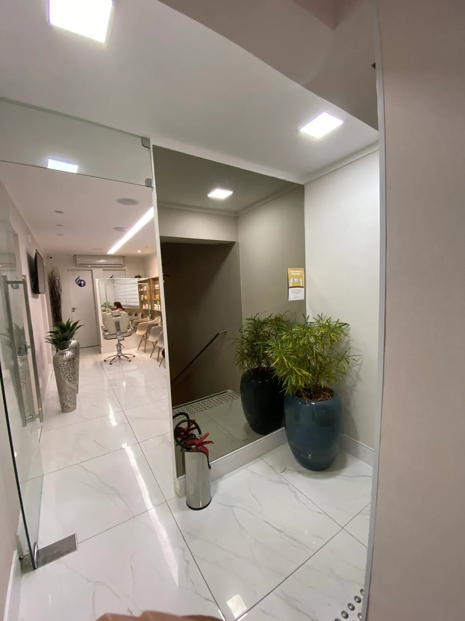 Circulação interna com espelho do piso ao teto ampliando o ambiente, vaso cerâmico vietnamita azul petróleo, piso tátil de acessibilidade e vista para os consultórios