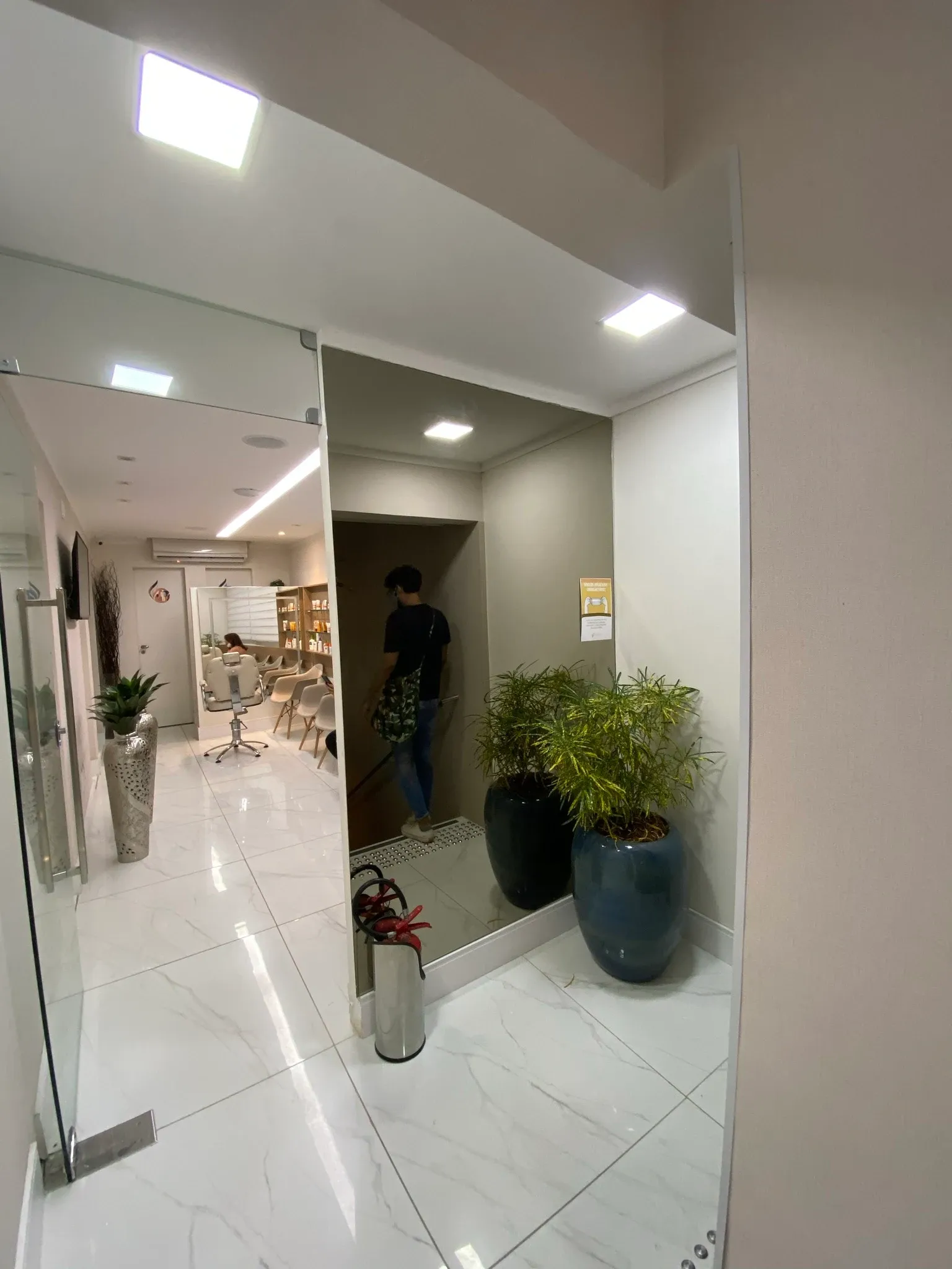 Transição entre os ambientes com paciente descendo a escadaria refletida no grande espelho vertical, piso claro e portas em vidro temperado