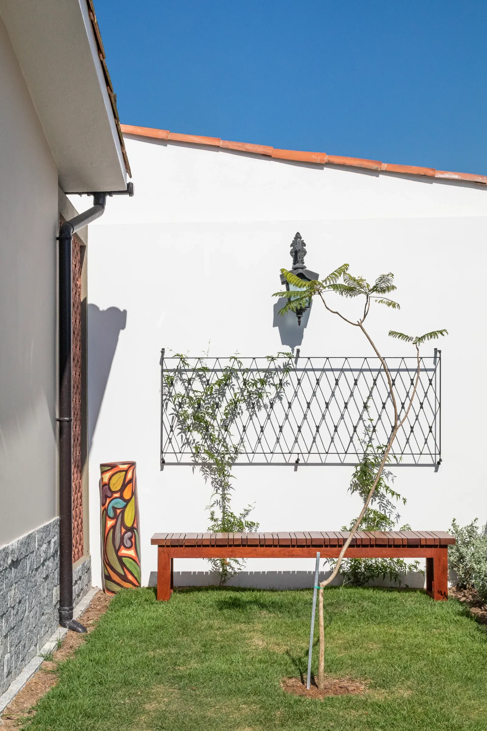 Pátio frontal ajardinado com banco em ripas de madeira maciça sob treliça metálica na parede caiada e vaso cerâmico artístico