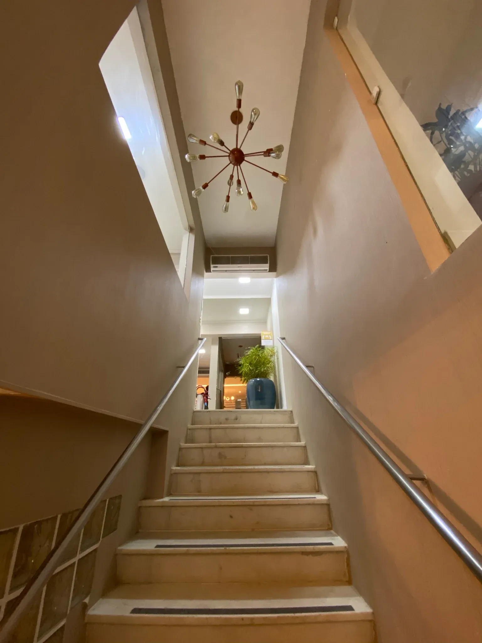 Escadaria em mármore claro com fitas antiderrapantes e corrimão duplo em aço inox, iluminada por lustre pendente Sputnik com lâmpadas aparentes de filamento