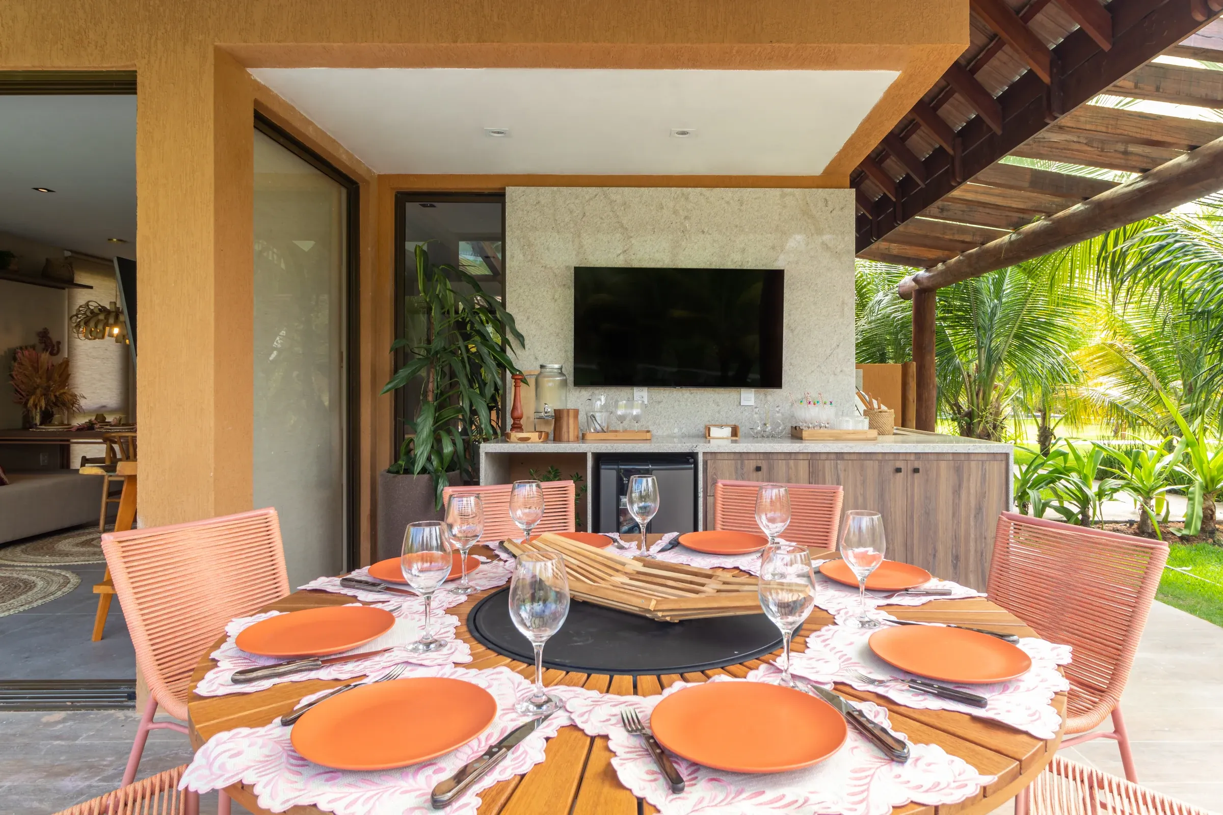 Varanda gourmet com mesa de jantar redonda e centro giratório com louça em terracota, bancada de apoio com TV e frigobar