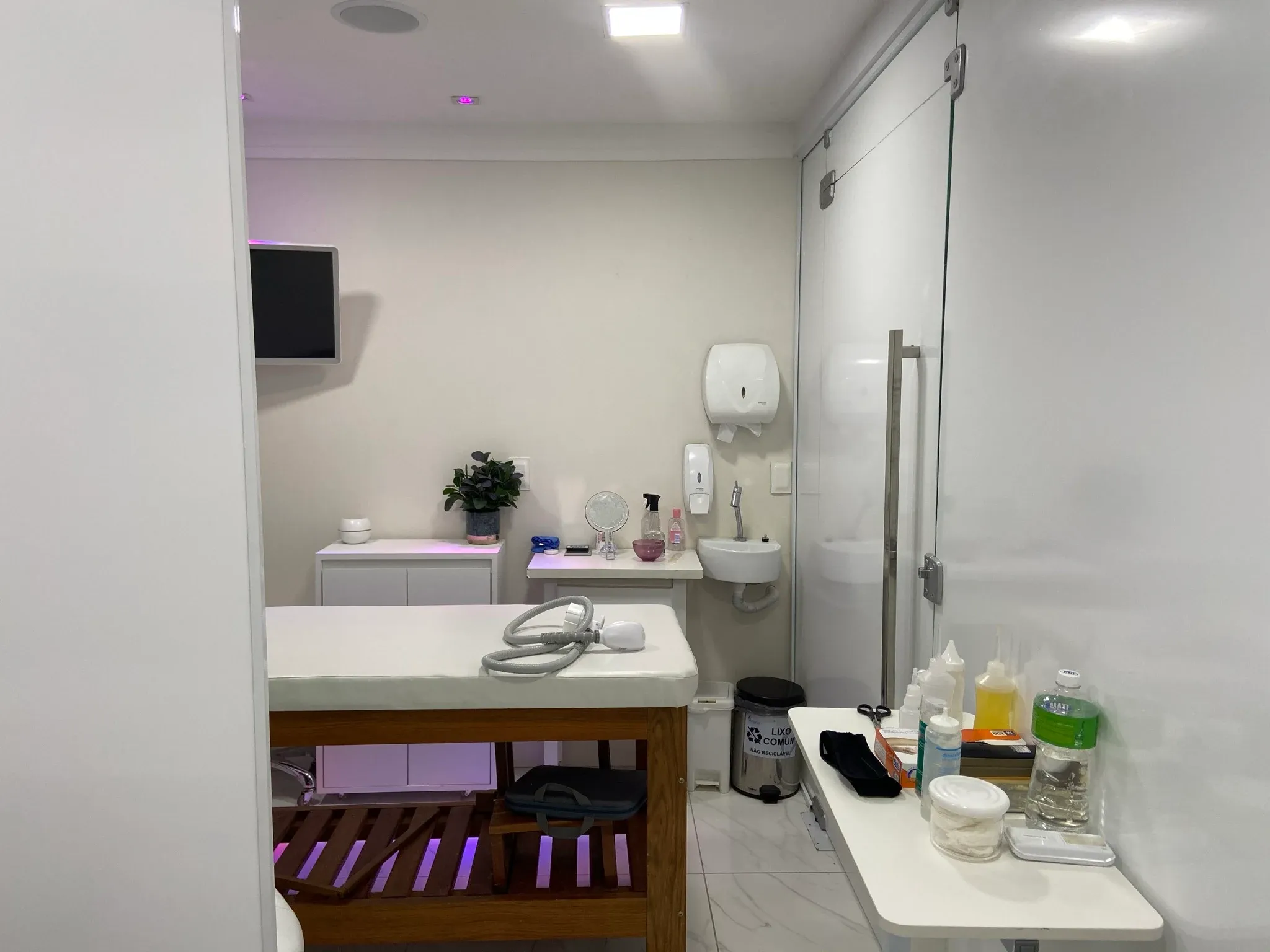 Sala de procedimentos estéticos com maca em madeira maciça acolchoada, bancada com pia para higienização asséptica, dispensadores hospitalares e iluminação indireta lilás