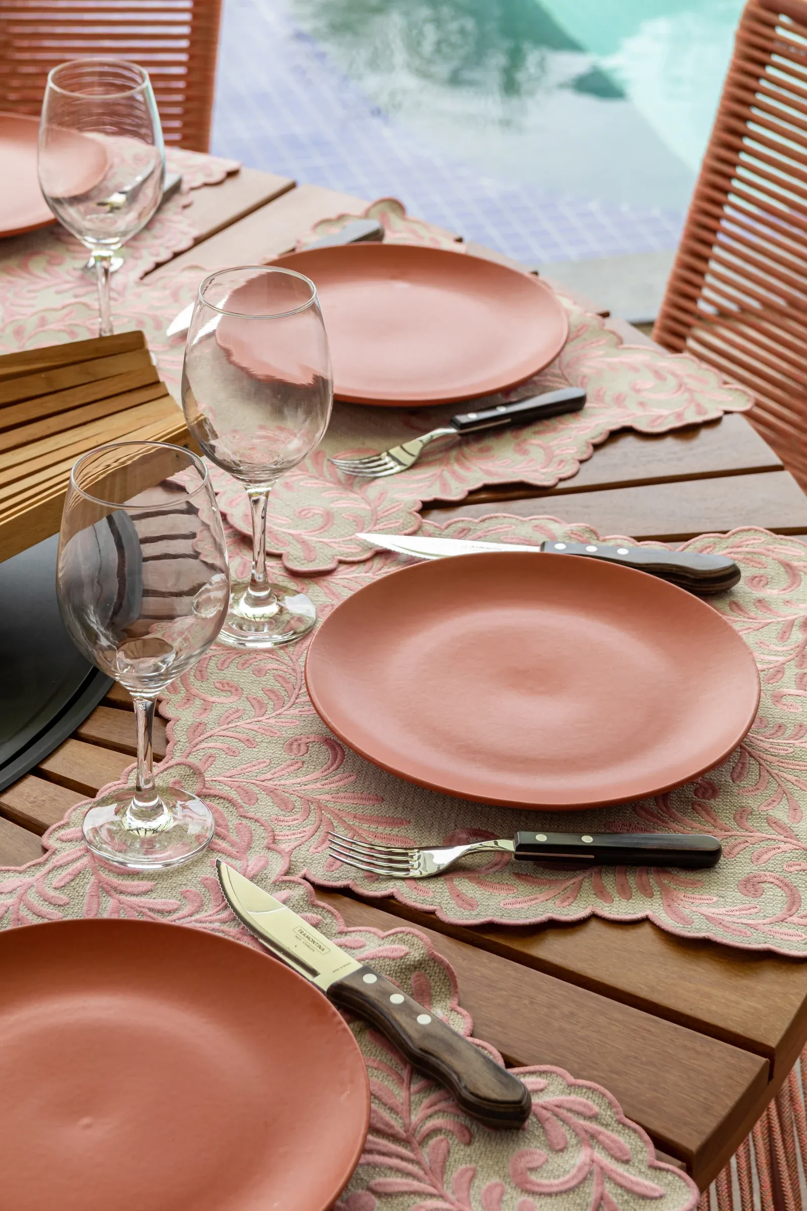 Detalhe da mesa posta com pratos cerâmicos em terracota, jogos americanos rendados rosé e taças de vinho