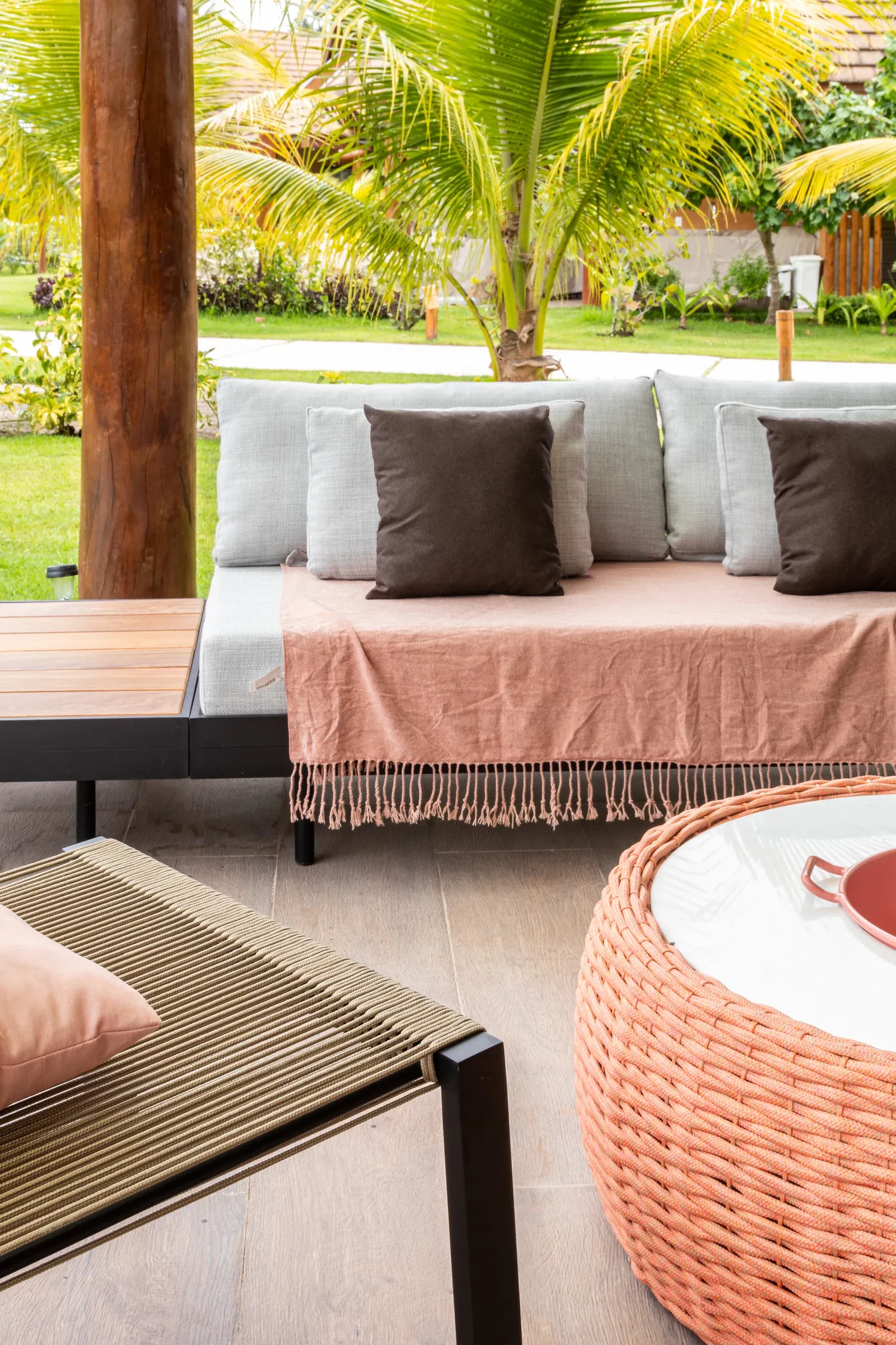 Lounge na varanda com estofado cinza, manta terracota com franjas artesanais, poltrona em corda náutica e mesa de centro redonda