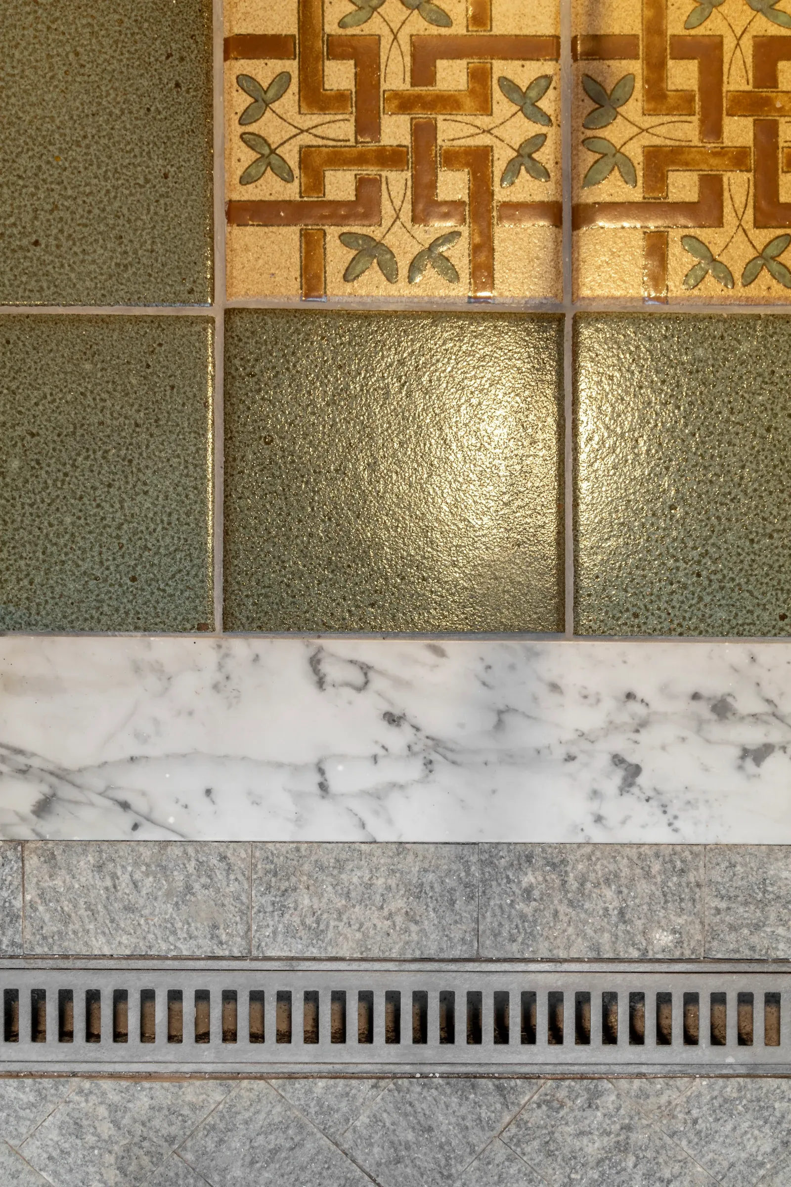 Encontro refinado de materiais no piso: ladrilho hidráulico floral clássico, cerâmica verde oliva, soleira em mármore branco e ralo linear em granito